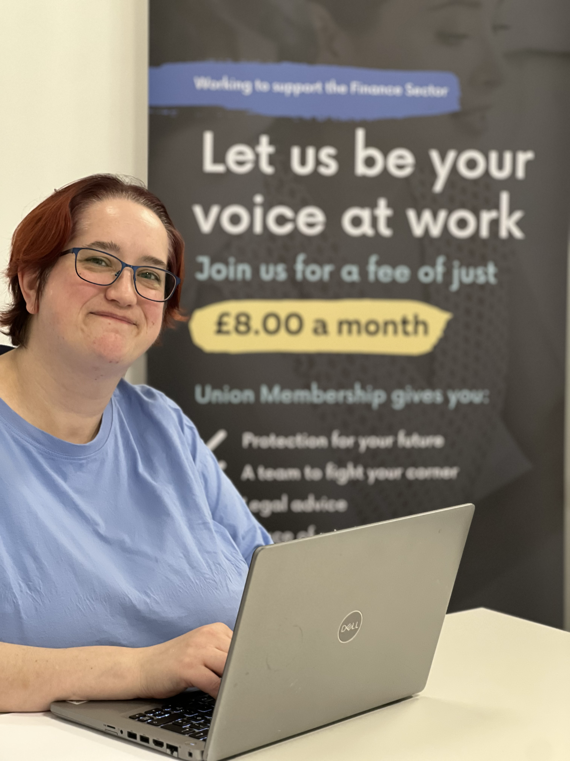 Smiling woman with a laptop sits in front of a banner saying 'Let us be your voice at work'
